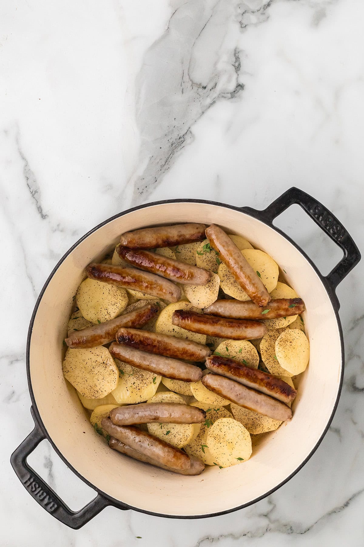 Sausages and sliced potatoes arranged in a white Dutch oven on a marble countertop.