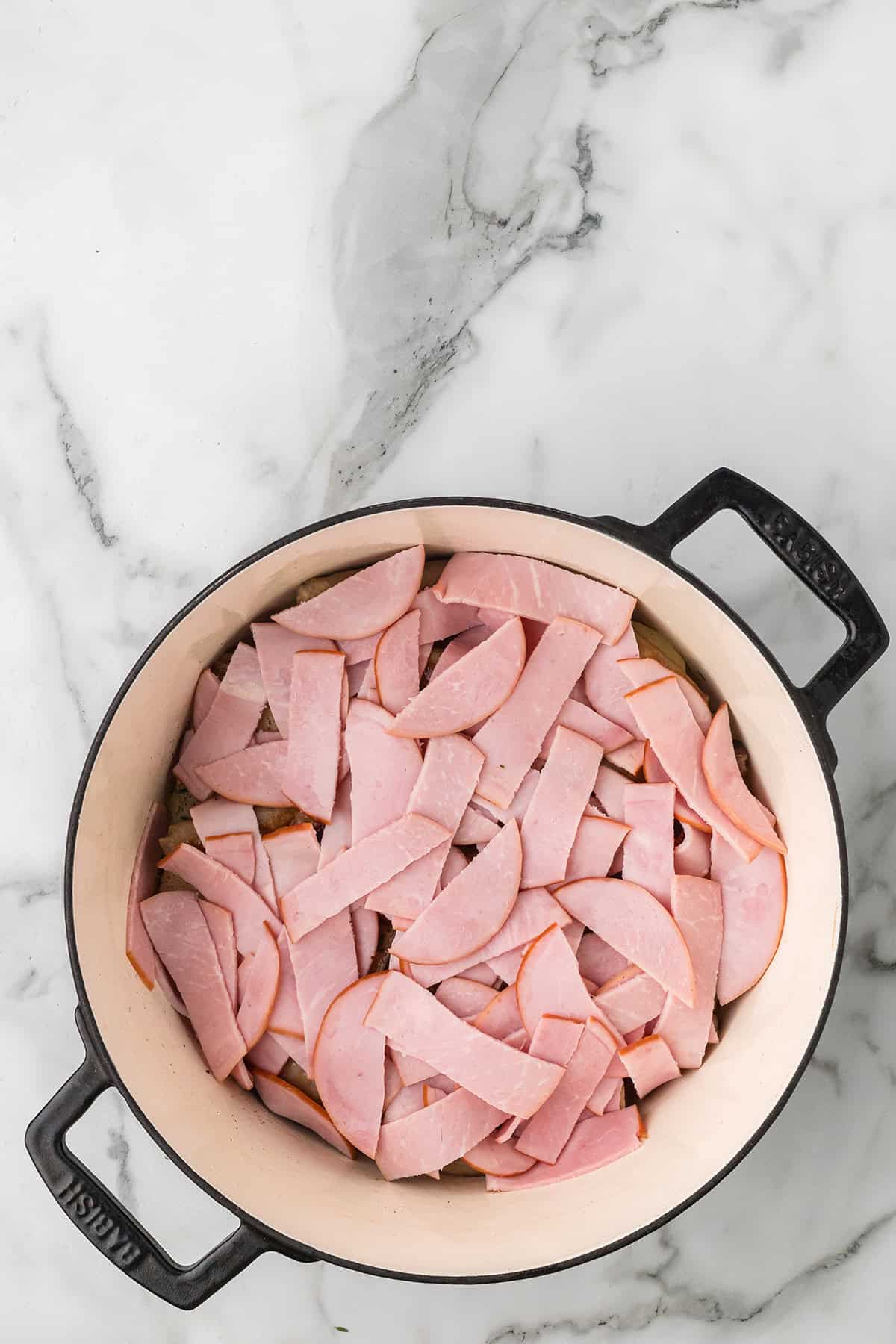 Sliced Canadian bacon pieces arranged in a white Dutch oven on a marble countertop.