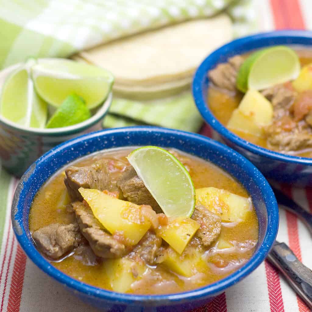 Two blue bowls filled with mexican beef stew (caldillo) with lime wedges and tortillas on the side.