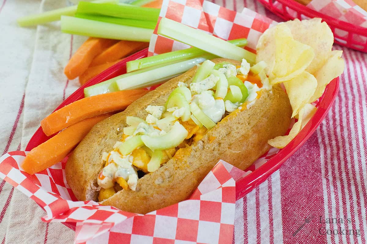 A hot dog bun filled with chopped vegetables, topped with dressing, served with potato chips, carrot sticks, and celery sticks in a red basket on a checkered paper.