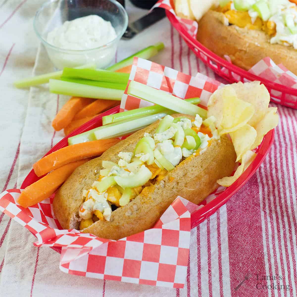 A hot dog bun filled with diced chicken, celery, and crumbled blue cheese, served with carrot and celery sticks, potato chips, and a cup of dipping sauce in a red basket.