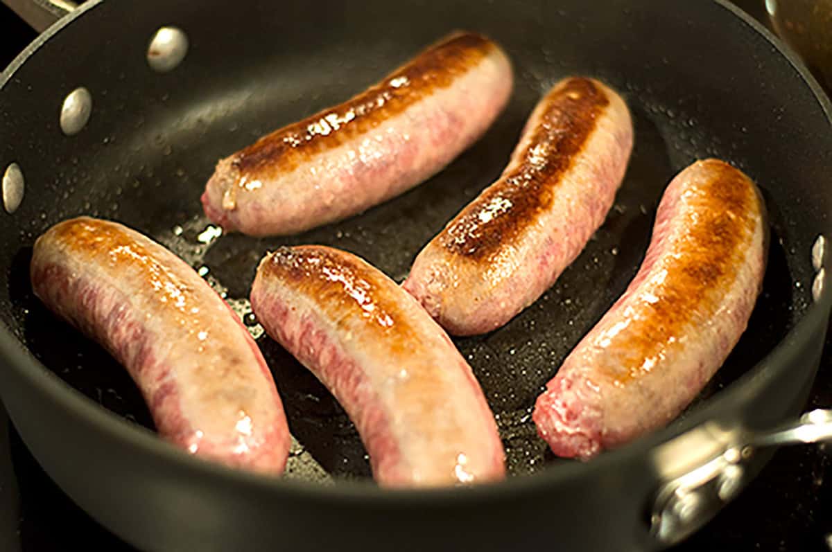 Five sausages are browning in a black frying pan on a stove, with some areas cooked and others still pink.
