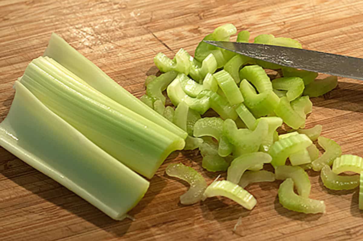 A knife slicing celery on a wooden cutting board, with whole and chopped celery pieces visible.
