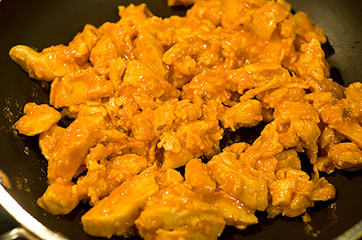 Cooked chicken pieces coated in Buffalo chicken sauce on a skillet.