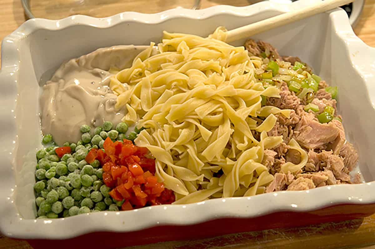Uncooked casserole ingredients: noodles, peas, red peppers, tuna, celery, and creamy sauce in a baking dish.