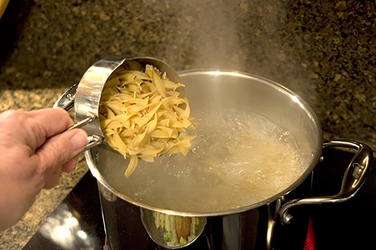 A hand pours dry pasta from a metal measuring cup into a pot of boiling water on a stovetop.