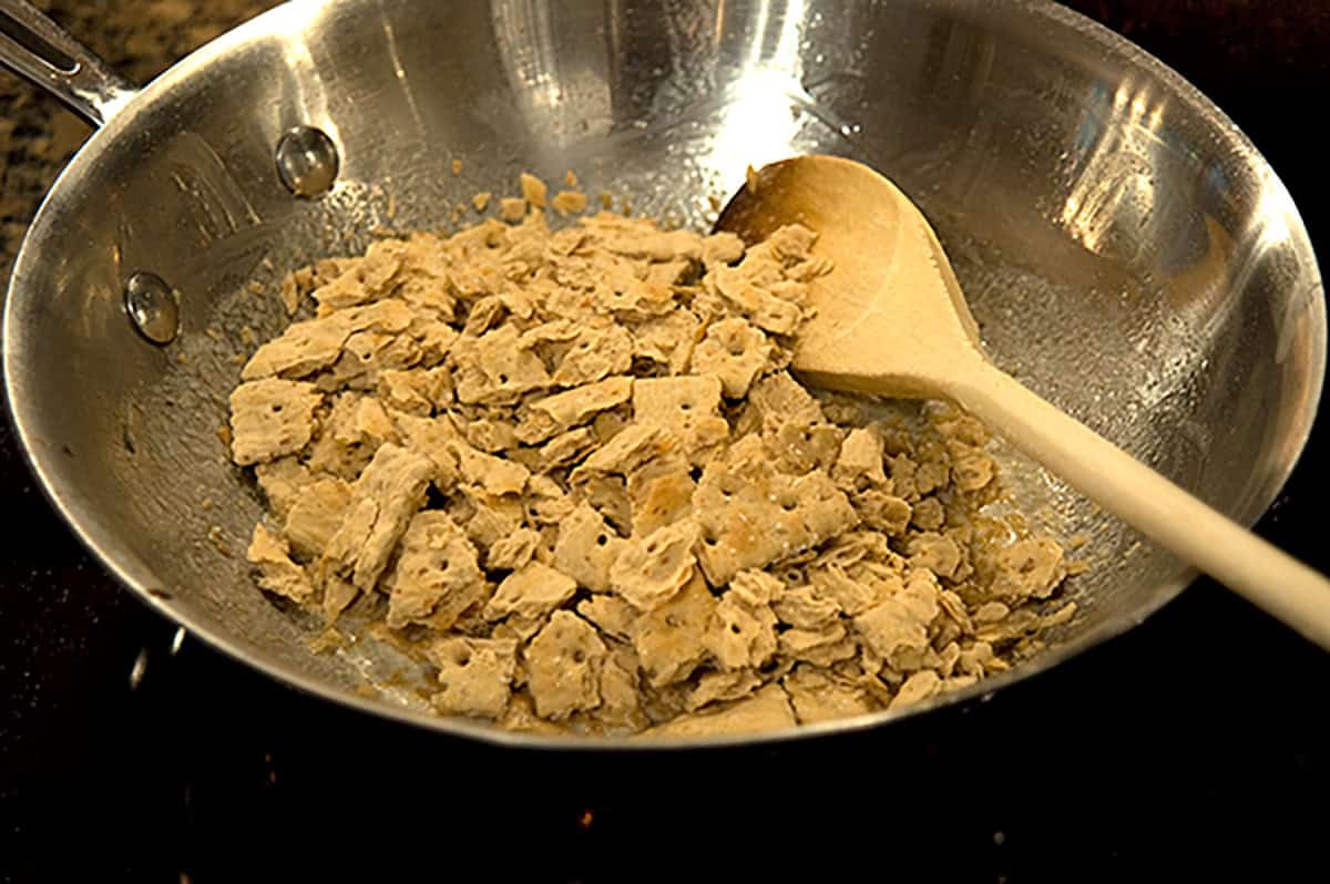 Broken cracker pieces being stirred in a metal pan with a wooden spoon.