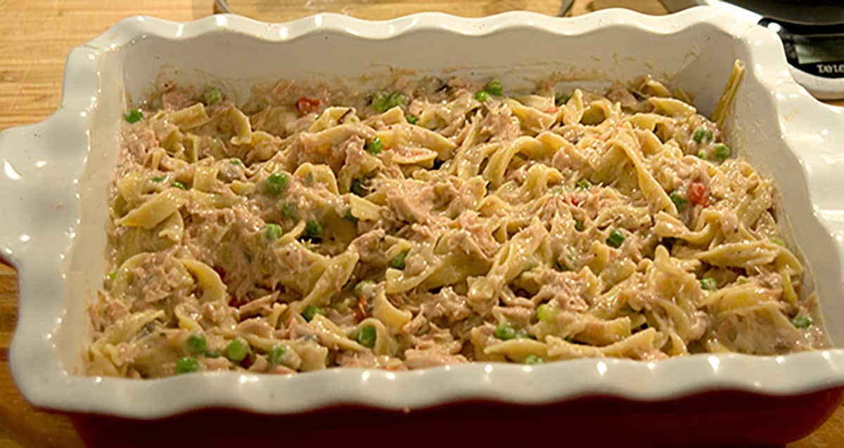 Baked tuna noodle casserole with peas in a white and red ceramic dish on a wooden surface.