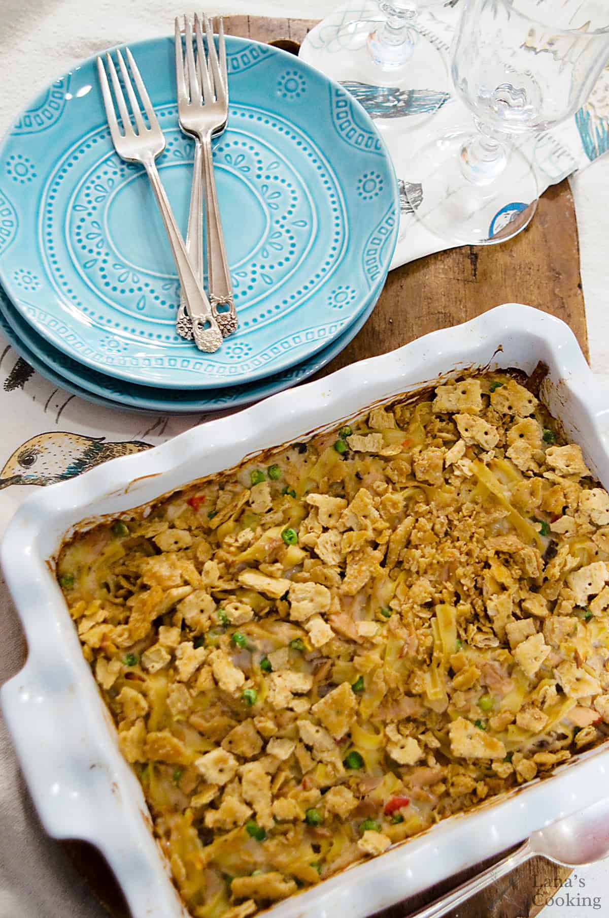 A baked casserole in a white dish next to stacked blue plates and three forks on a wooden surface.