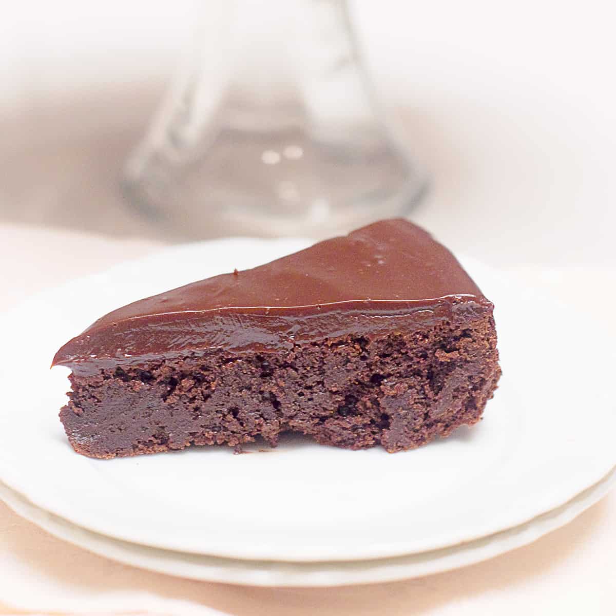 A serving of flourless chocolate cake on a white plate with remainder of cake on a cake stand in background.