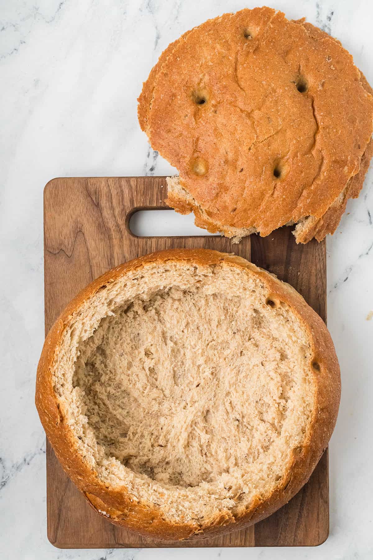 Round loaf of bread with the top cut off and the inside hollowed out, on a wooden cutting board.