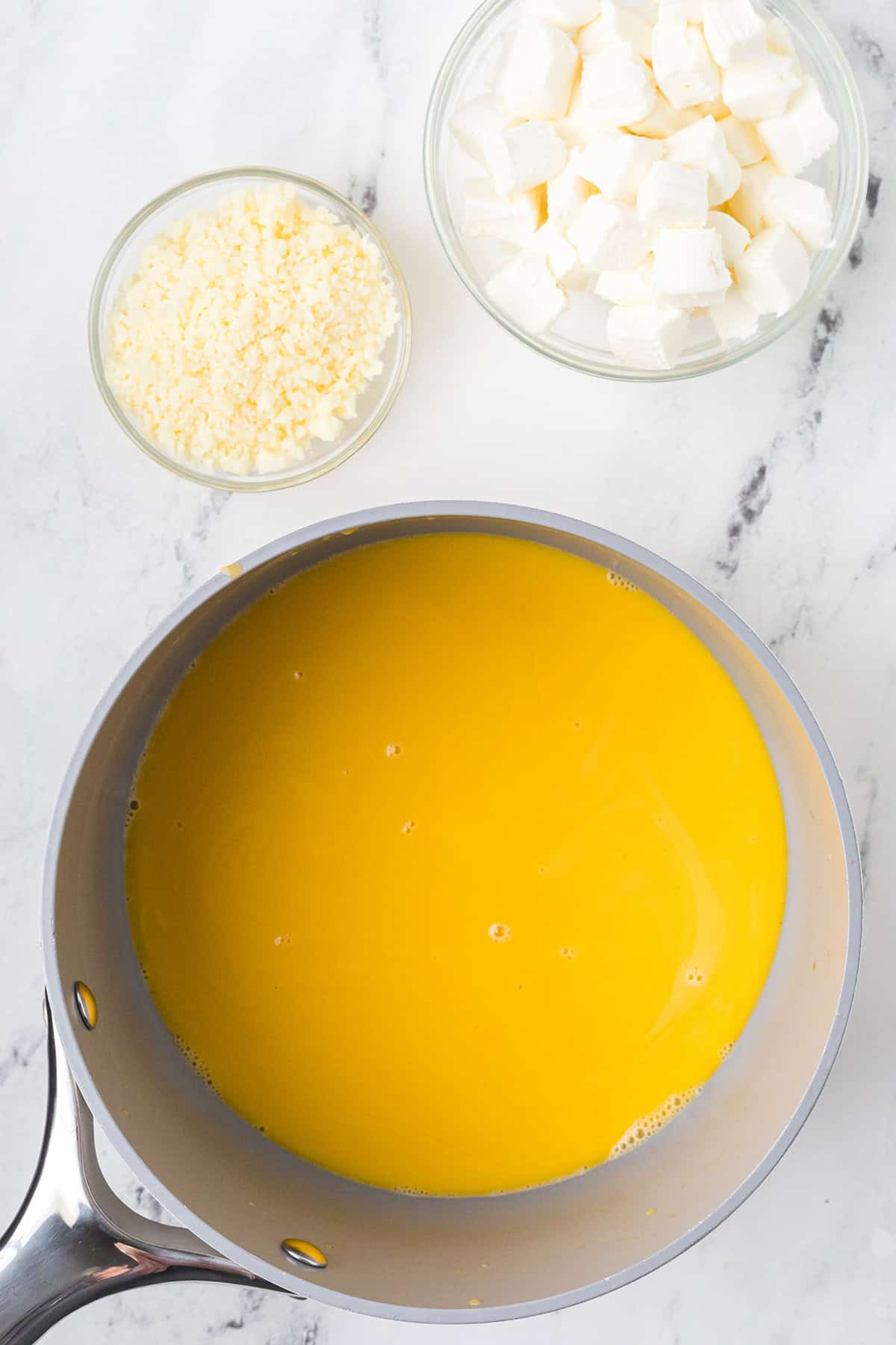 A saucepan with melted cheddar cheese, next to bowls of white cubes and grated cheese, on a marble counter.