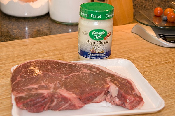 Raw steak on a foam tray and a jar of bleu cheese dressing on a wooden counter.