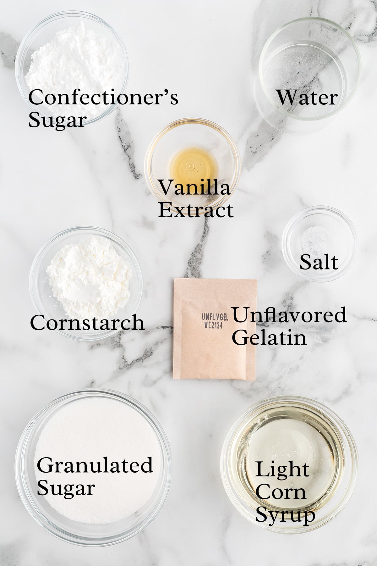 Bowls of sugar, cornstarch, salt, and other ingredients for homemade marshmallows arranged on a marble surface.