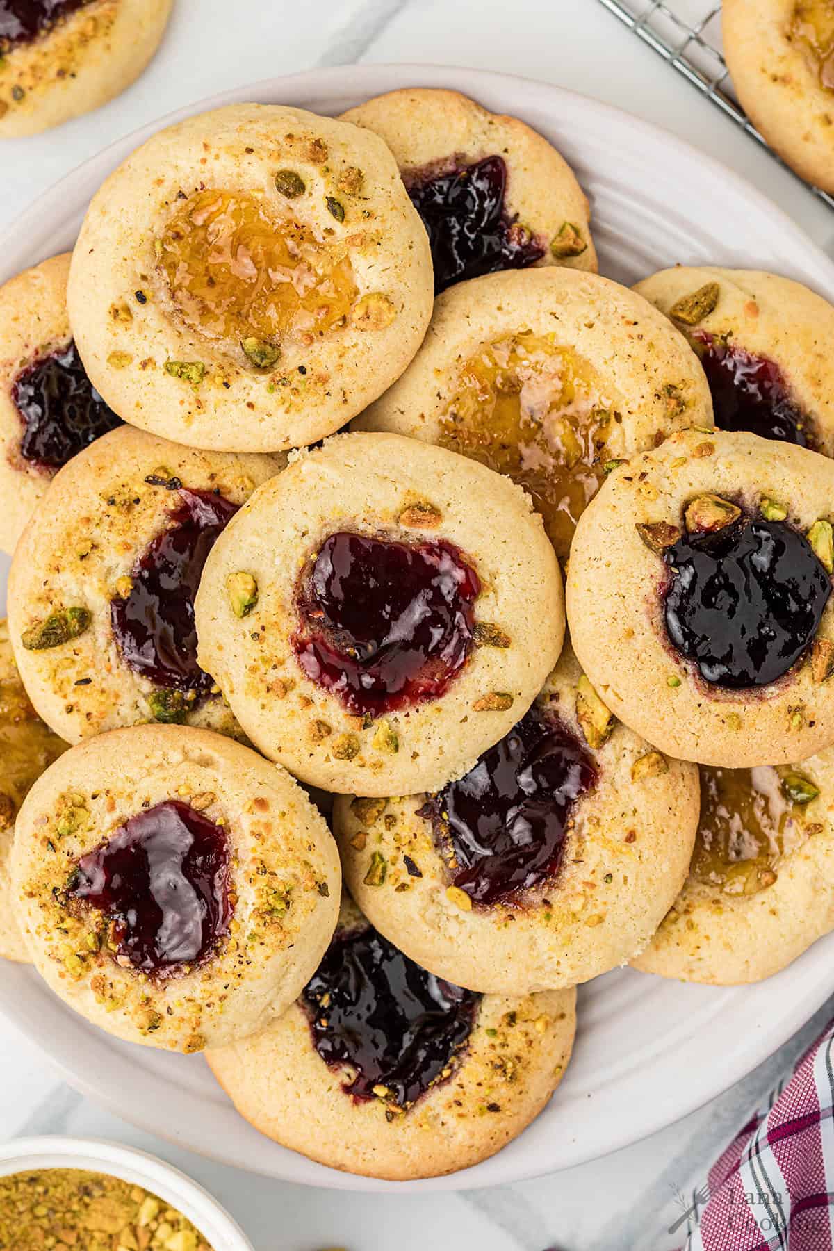 A plate of thumbprint cookies filled with red and yellow jam, topped with chopped pistachios.