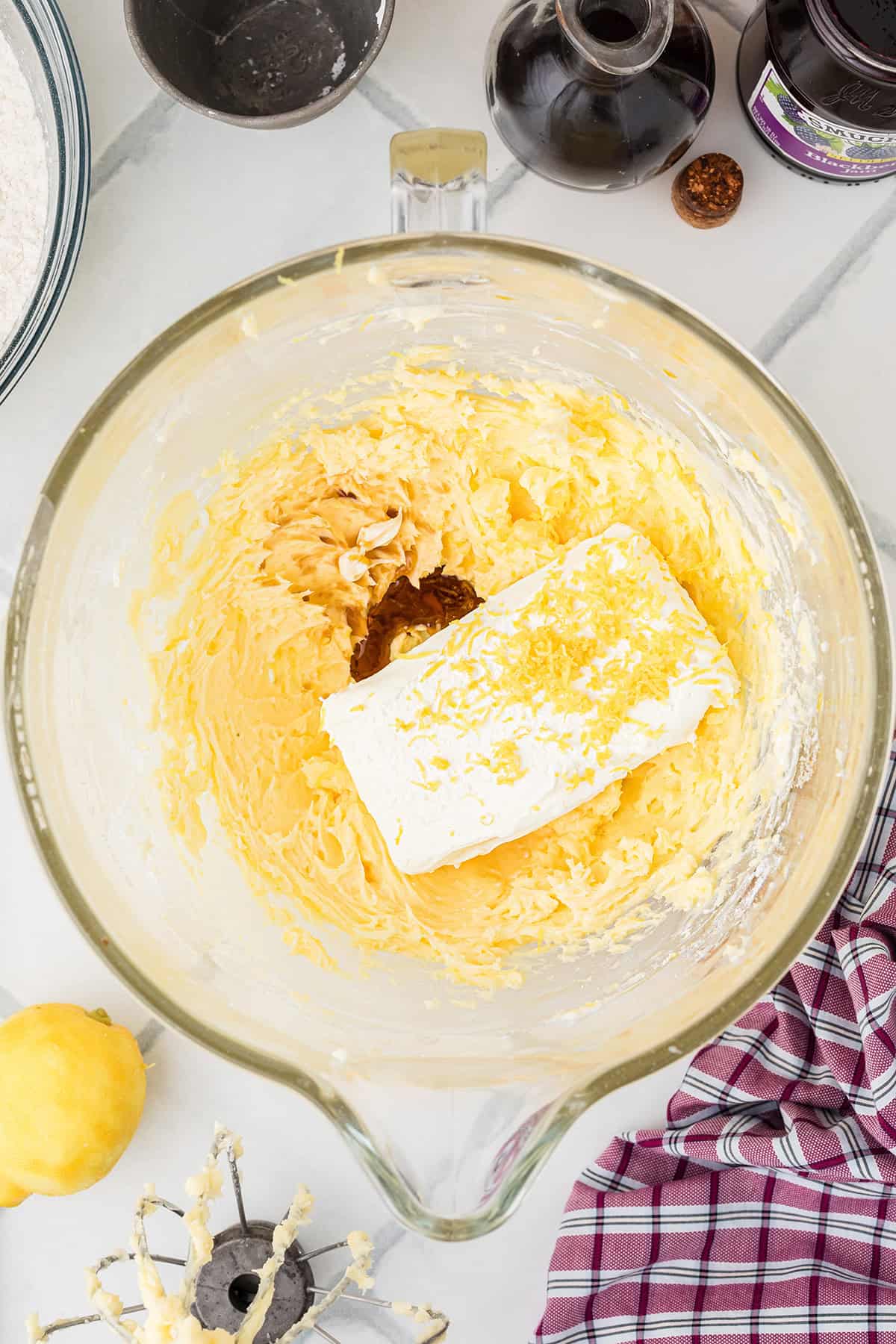 Mixer bowl with creamed batter, block of cream cheese, and lemon zest on top, surrounded by baking ingredients.