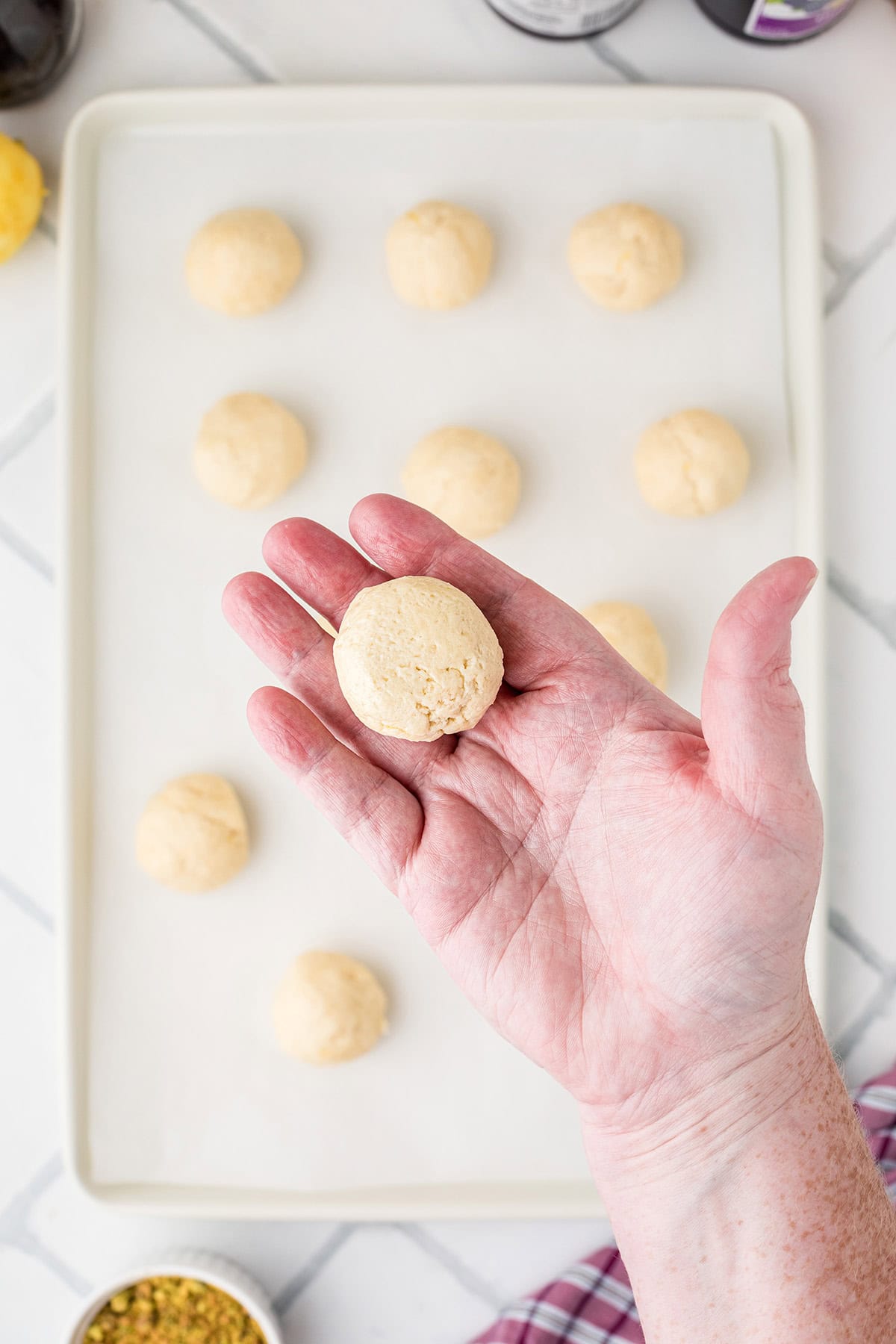 A hand holds a round ball of cookie dough above a baking sheet with more dough balls.