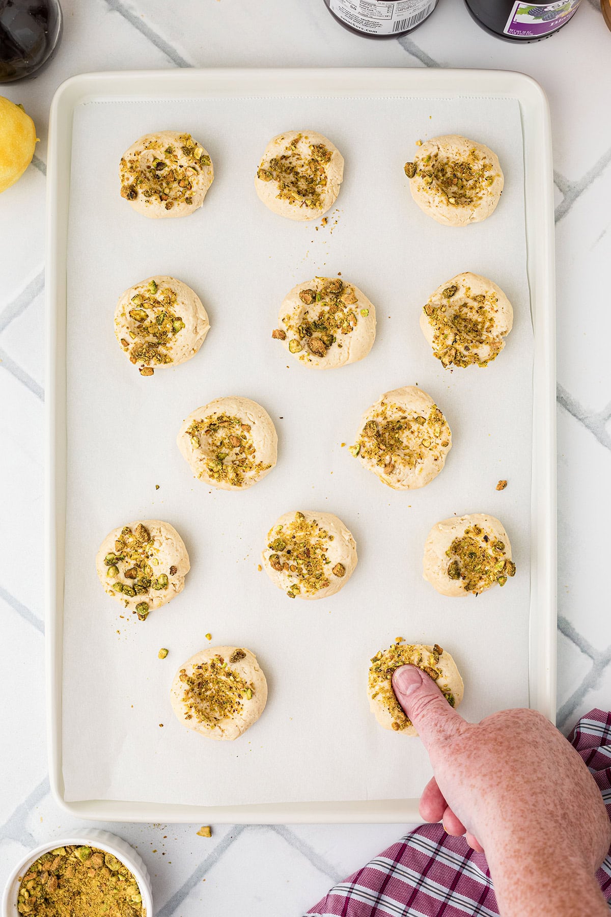 A hand presses a cookie on a baking tray with unbaked cookies topped with crushed pistachios.