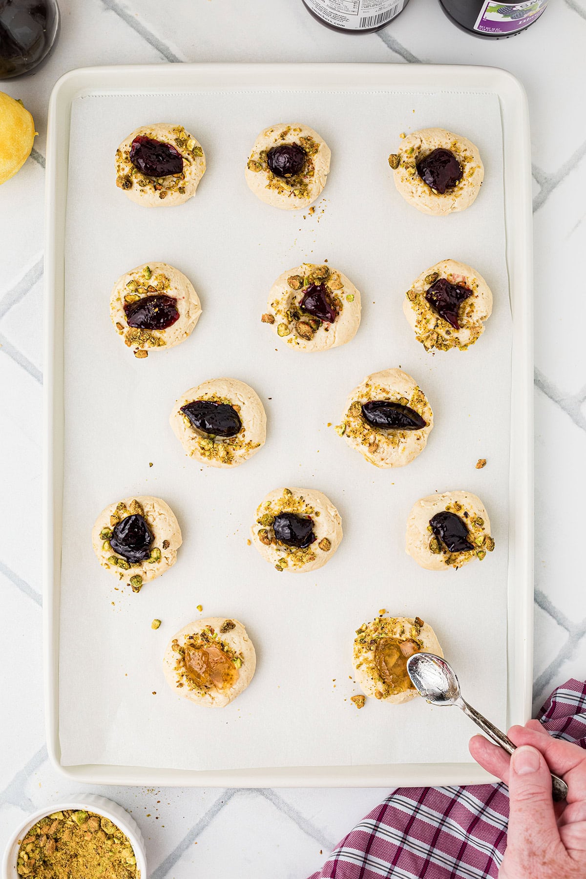A baking sheet with cookies topped with jam and chopped pistachios, with a hand spooning jam onto one cookie.