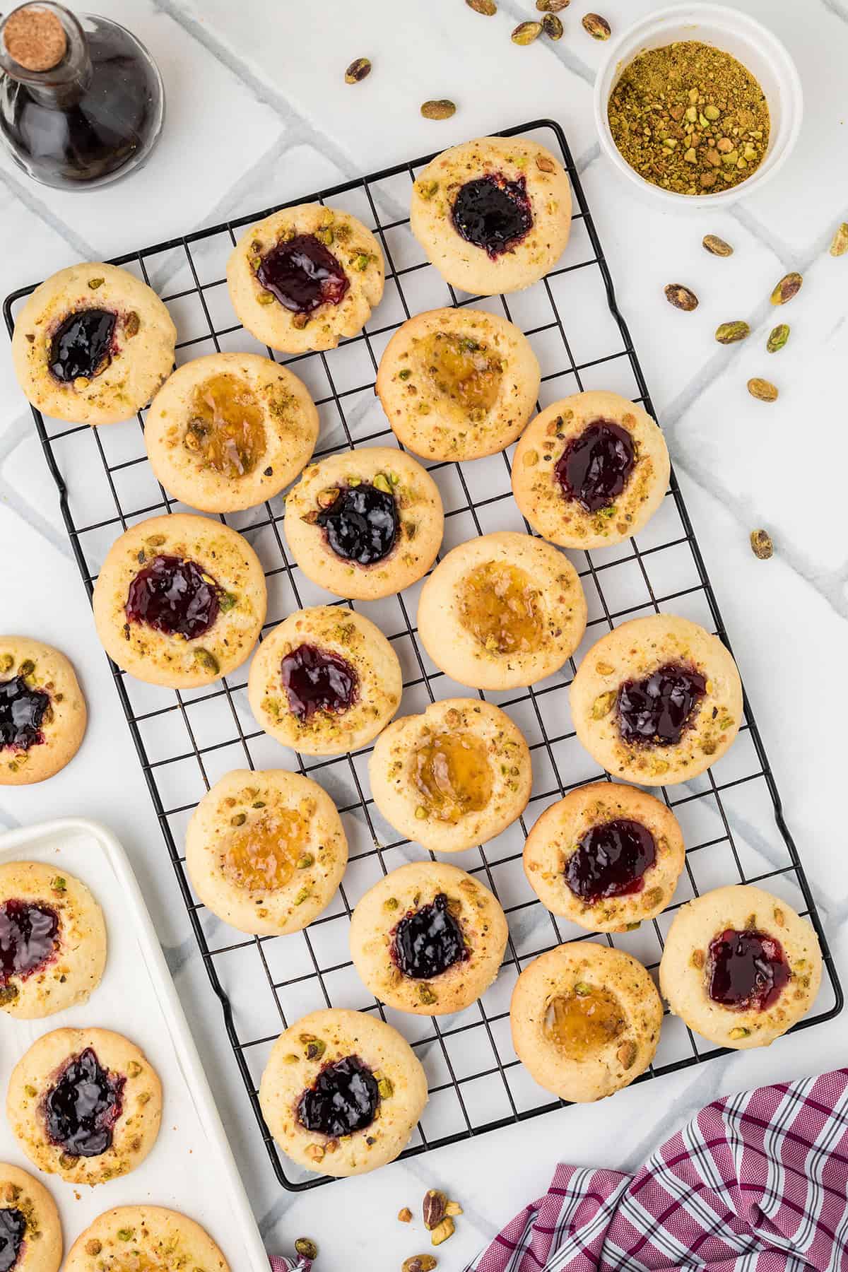 Jam-filled thumbprint cookies on a cooling rack, topped with pistachios, with scattered pistachios around.
