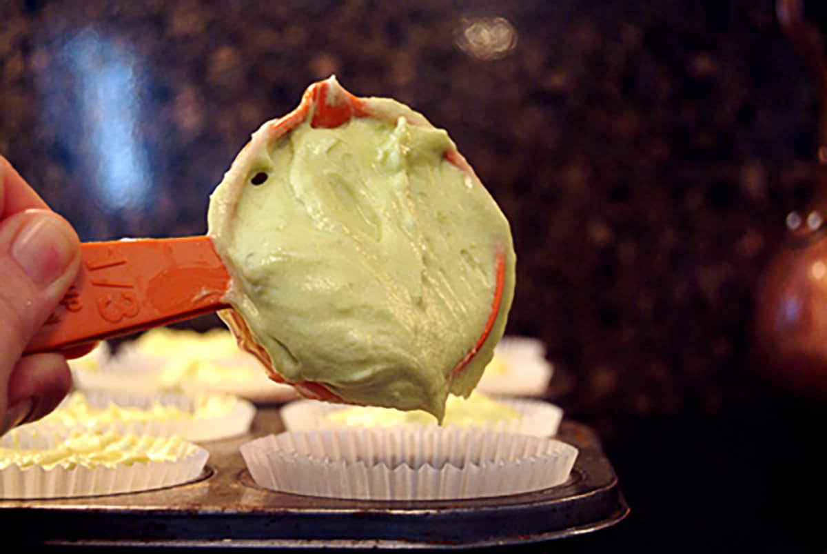 A spoon with green batter held above a muffin tin lined with paper cupcake liners.