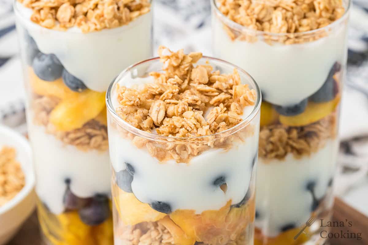 Three glasses filled with layers of granola, yogurt, blueberries, and peach slices, viewed from above.