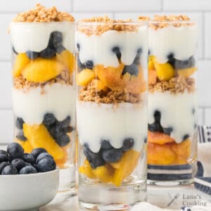 Three tall glasses layered with yogurt, peaches, blueberries, and granola, with a bowl of blueberries nearby.