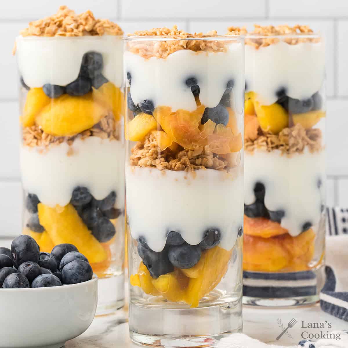 Three tall glasses layered with yogurt, peaches, blueberries, and granola, with a bowl of blueberries nearby.