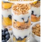 A glass parfait layered with yogurt, peaches, blueberries, and granola, with fresh blueberries and granola nearby.