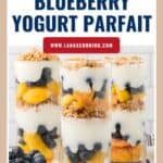 Three glasses filled with layers of yogurt, peaches, blueberries, and granola, with fresh blueberries beside them.