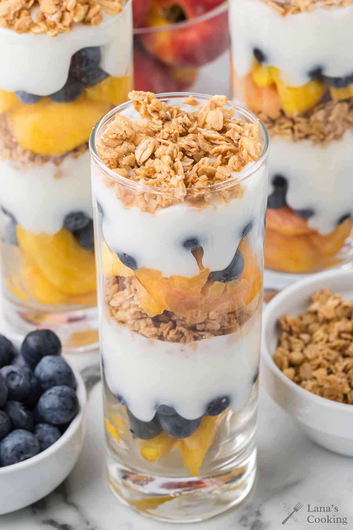 A glass filled with layered yogurt, granola, blueberries, and peaches, surrounded by fruit and granola bowls.