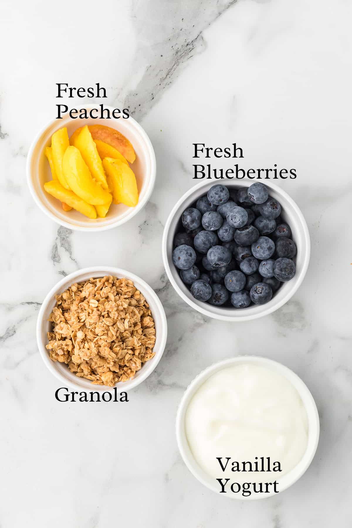 Four bowls containing fresh peach slices, blueberries, granola, and vanilla yogurt on a white surface.