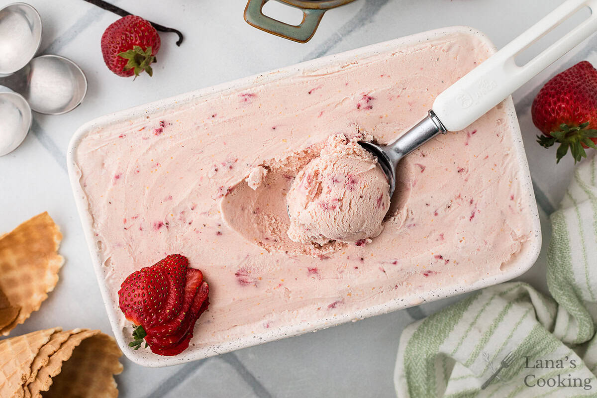 A rectangular container of strawberry ice cream with a scoop taken out, an ice cream scooper inside, and fresh strawberries and waffle cones nearby.