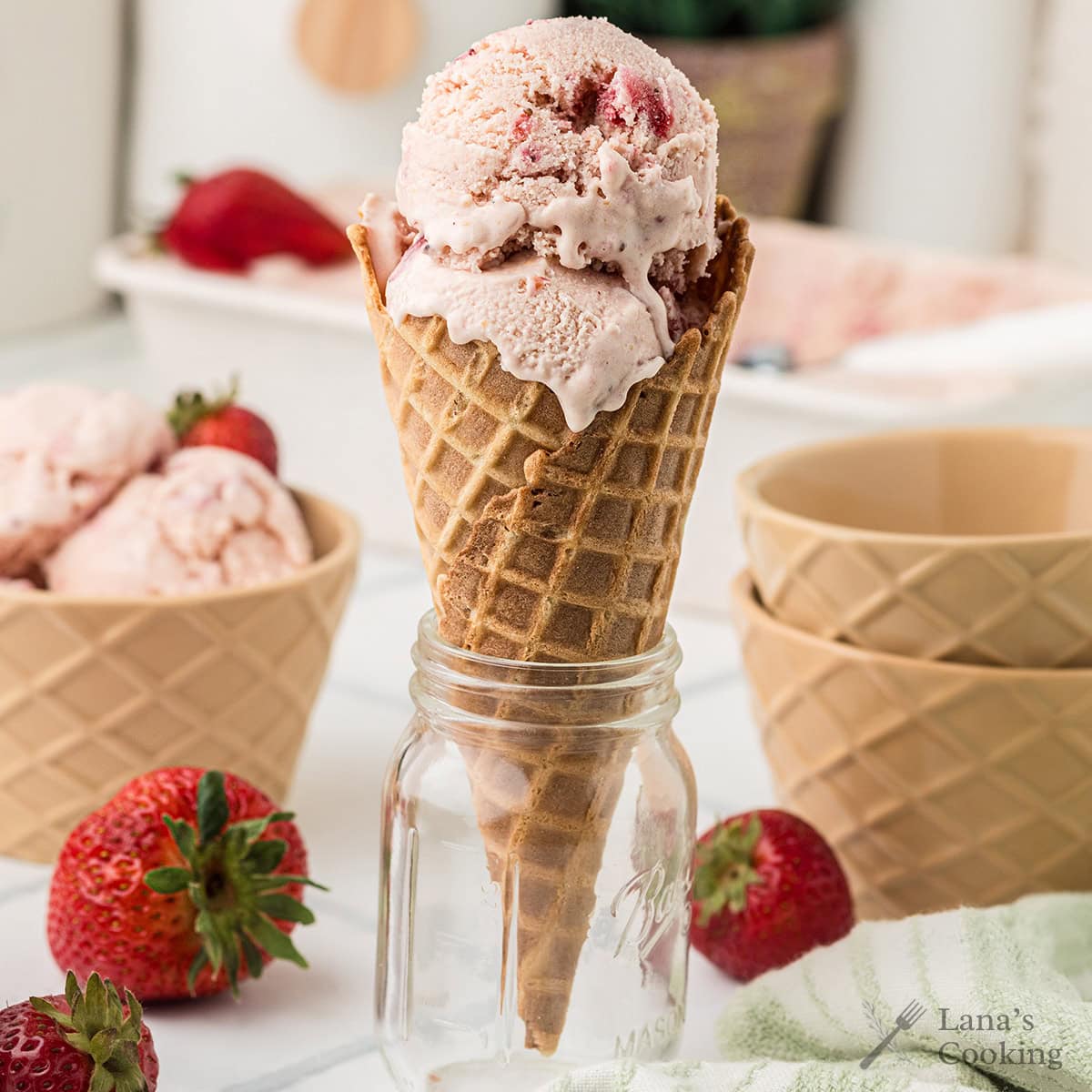 A waffle cone filled with strawberry ice cream sits upright in a glass jar, surrounded by fresh strawberries and empty bowls.