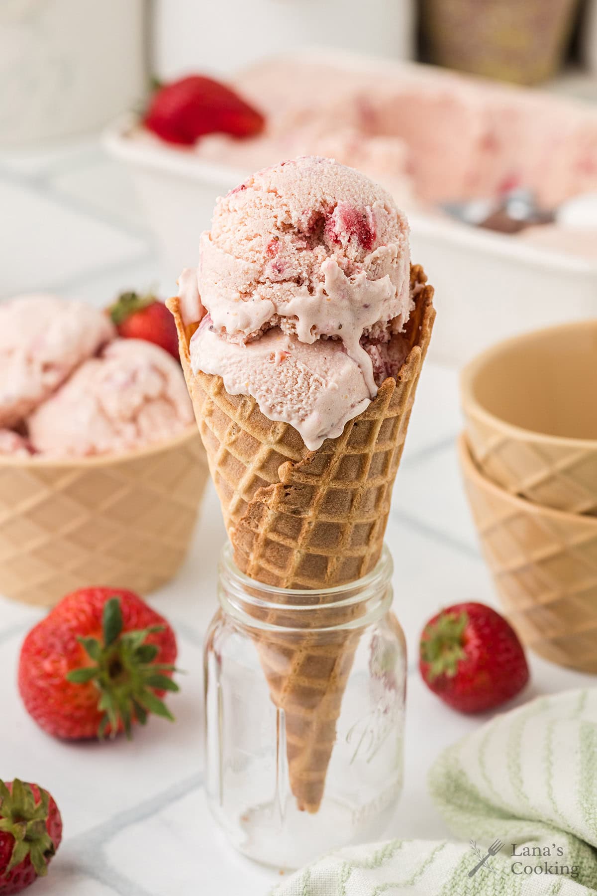 A waffle cone filled with two scoops of strawberry ice cream stands upright in a glass jar, with fresh strawberries and empty cones in the background.