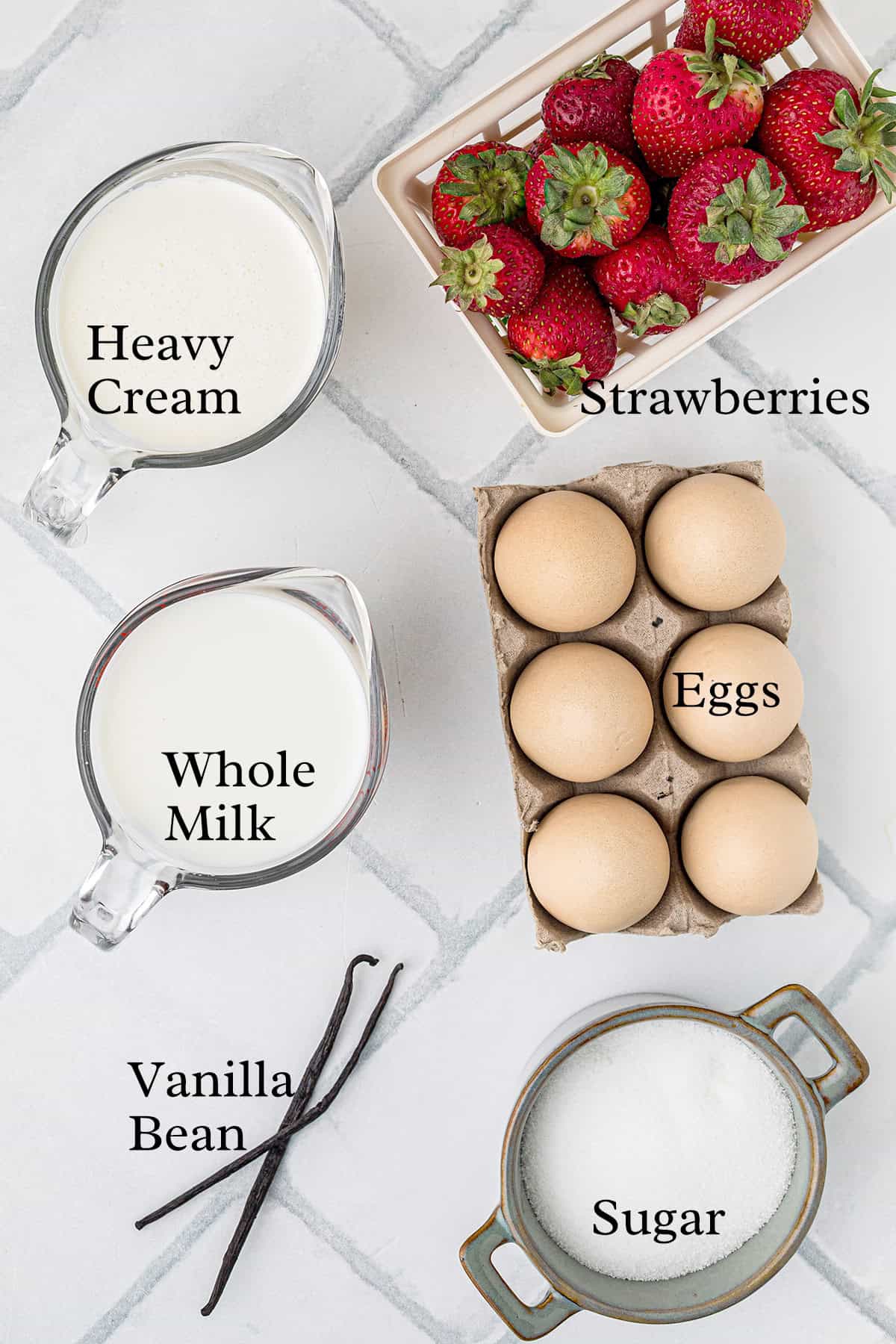 Overhead view of labeled ingredients for the recipe: heavy cream, whole milk, strawberries, eggs, vanilla bean, and sugar on a white tiled surface.