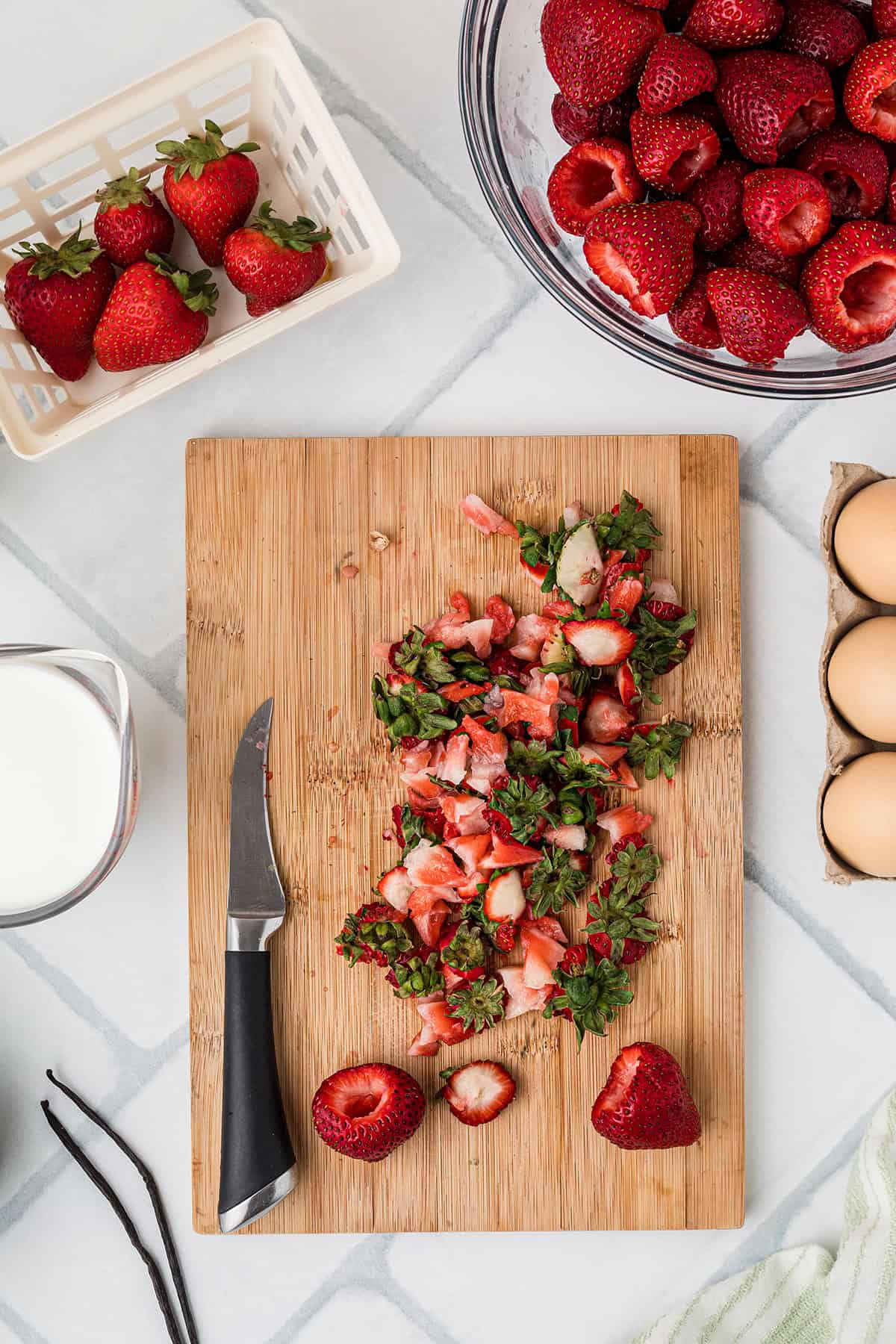 A wooden cutting board with strawberry tops and a paring knife, surrounded by whole strawberries, a bowl of hulled strawberries, eggs, milk, and a vanilla bean.
