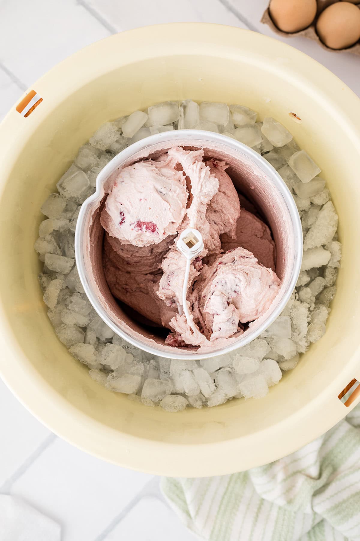 A close-up of a homemade ice cream maker with strawberry ice cream inside, surrounded by ice cubes in the outer bowl.