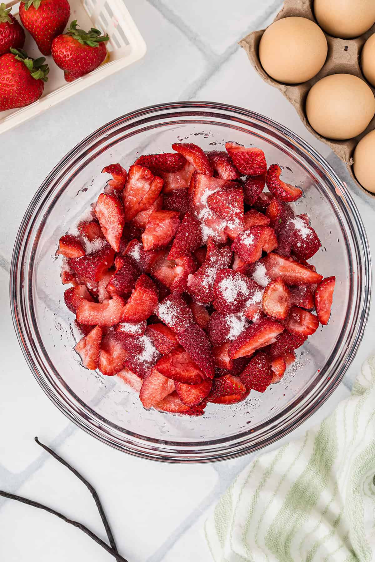 A glass bowl of sliced strawberries sprinkled with sugar on a white countertop, next to eggs, whole strawberries, a green-striped towel, and a vanilla bean.