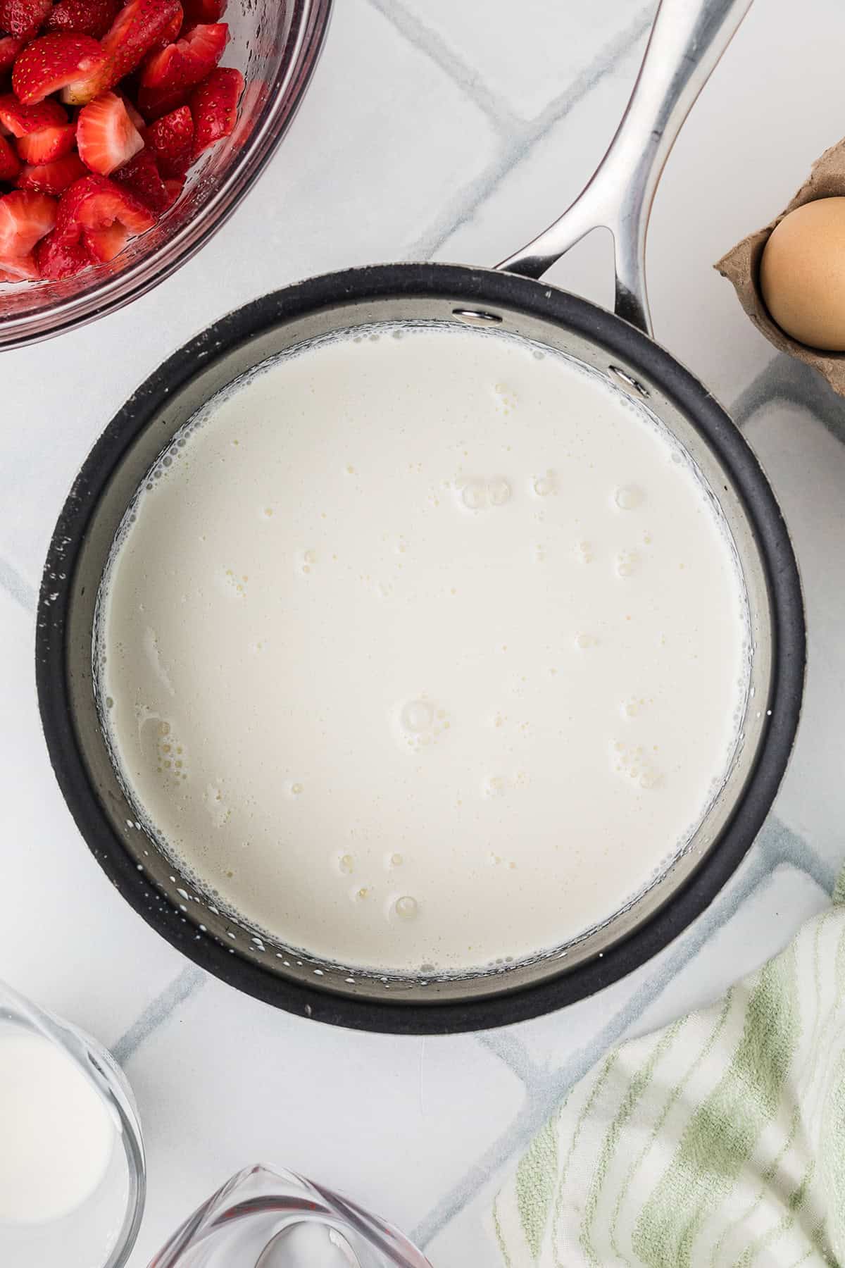 A saucepan filled with a creamy liquid sits on a countertop, surrounded by a bowl of sliced strawberries, eggs, and a striped towel.