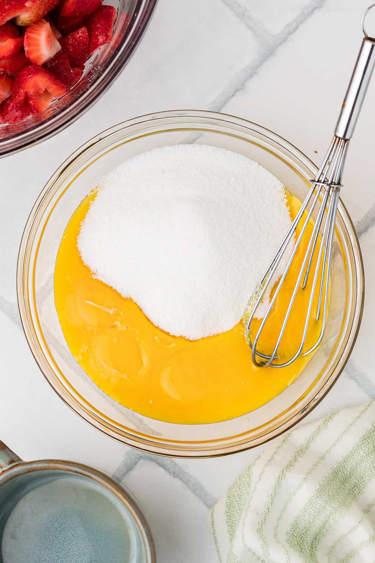 A glass bowl containing egg yolks and sugar with a whisk, next to a bowl of sliced strawberries and a striped towel on a white surface.