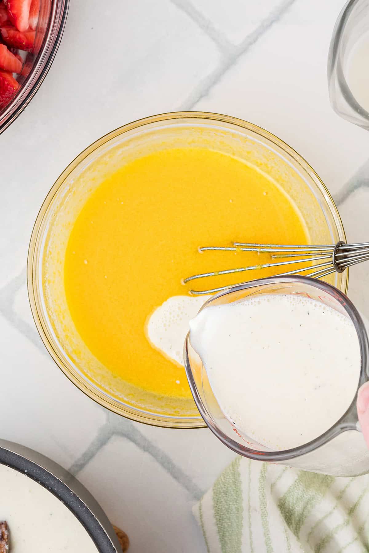 A hand pours a creamy liquid from a measuring cup into a bowl egg yolks and sugar with a whisk, on a white tiled surface.