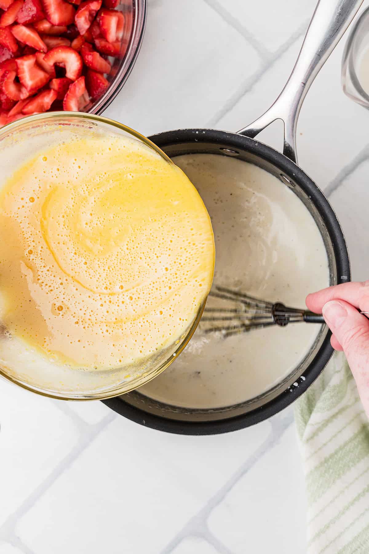 A bowl of egg-sugar mixture is being poured into a saucepan of cream mixture while a hand whisks the contents; a bowl of sliced strawberries sits nearby.