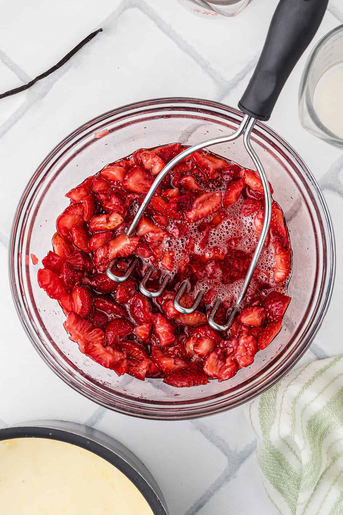 A glass bowl filled with crushed strawberries and a metal potato masher, sitting on a tiled countertop.