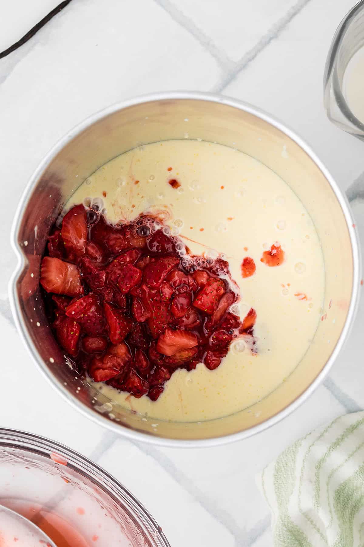 A stainless steel mixing bowl containing the ice cream custard base and chopped strawberries on a white surface.