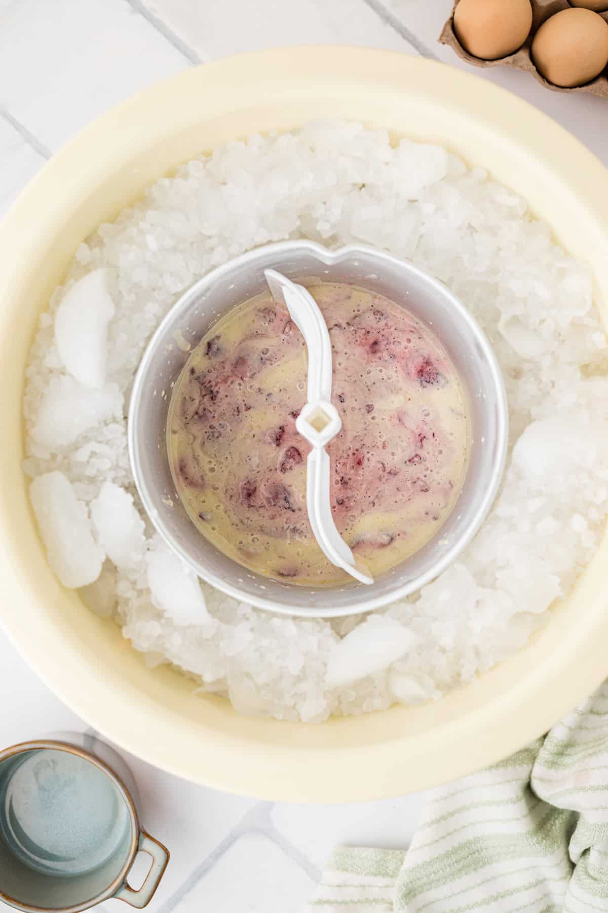 An ice cream maker with berry ice cream mixture surrounded by ice. Eggs, a striped towel, and a cup appear on a marble surface.