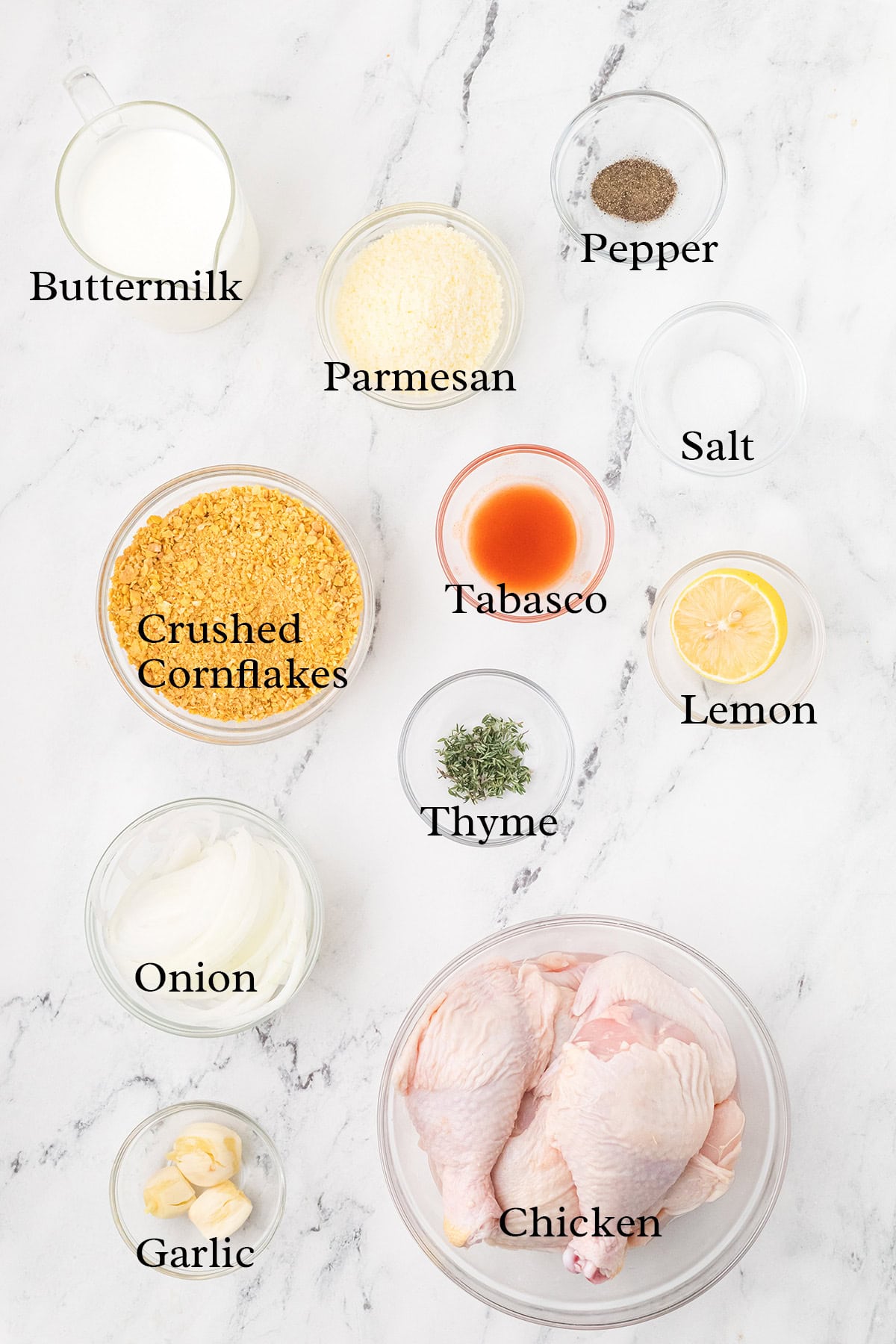 Bowls of buttermilk, parmesan, cornflakes, spices, onion, garlic, lemon, and raw chicken on a marble surface.