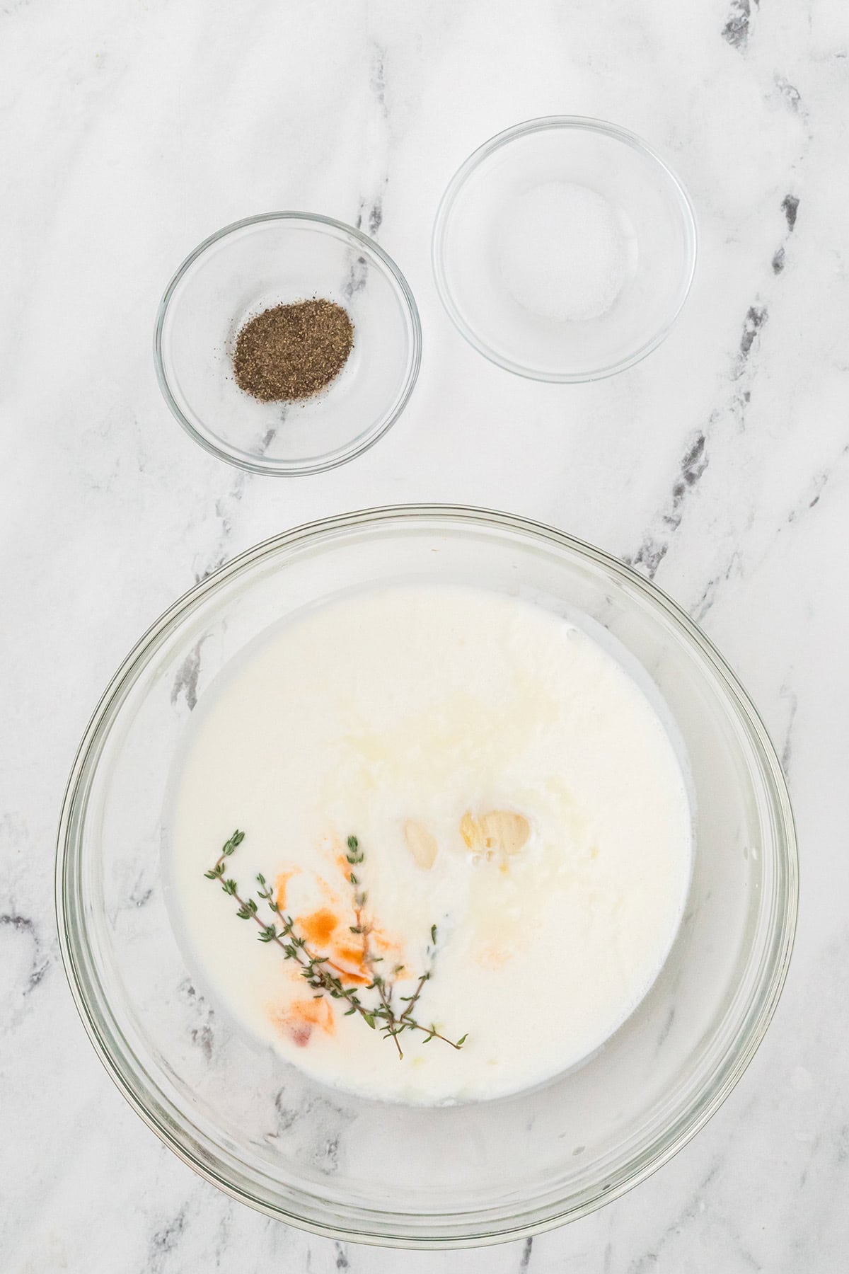 A bowl of buttermilk with herbs, garlic, and spices, plus two small bowls of black pepper and salt on marble.