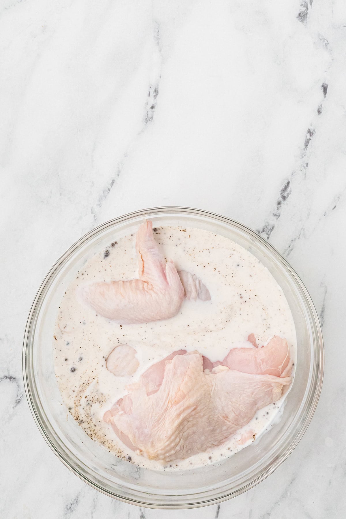 Raw chicken pieces in a bowl of marinade with black pepper, on a white marble surface.