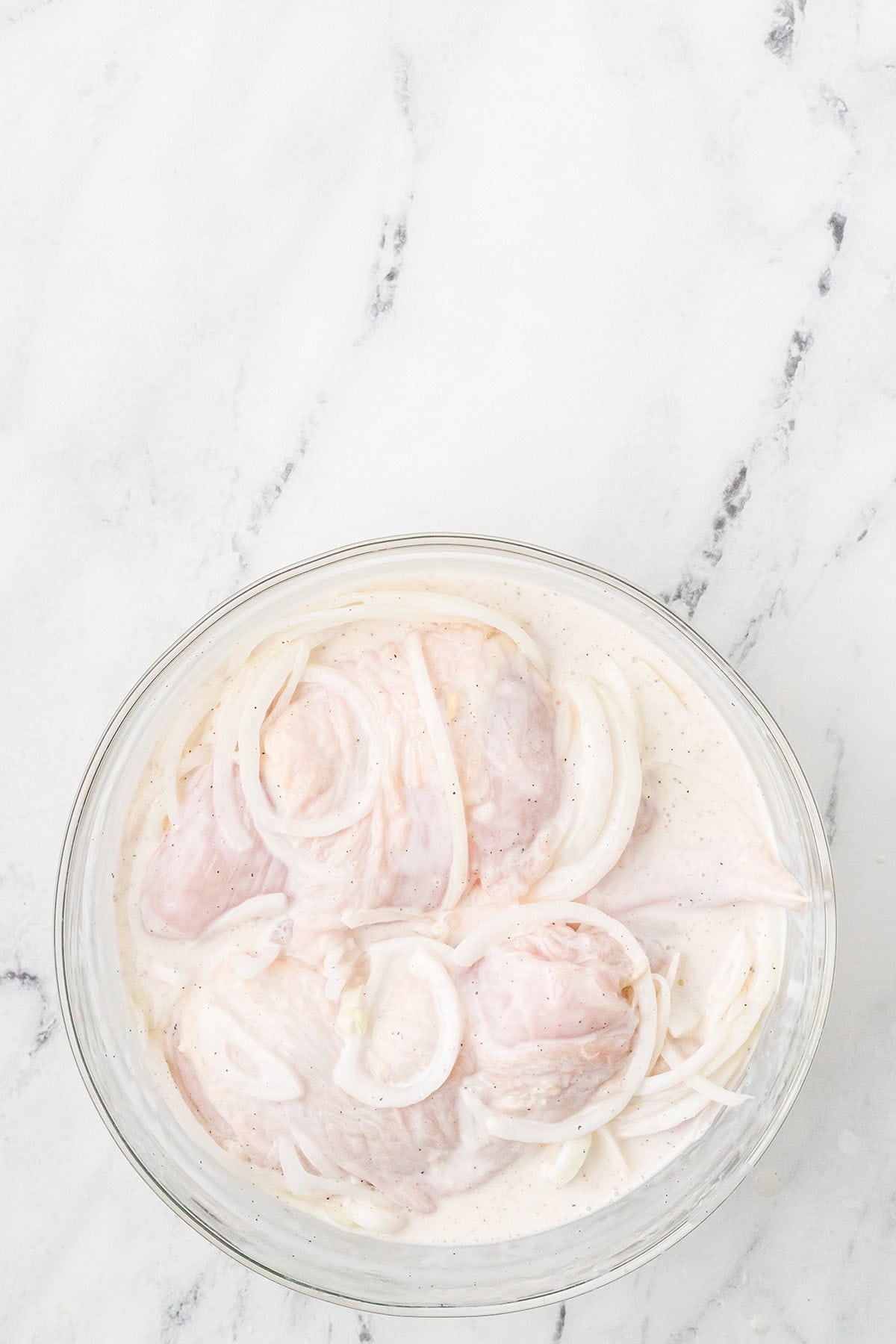 Raw chicken and onion slices marinating in buttermilk in a glass bowl on a marble surface.